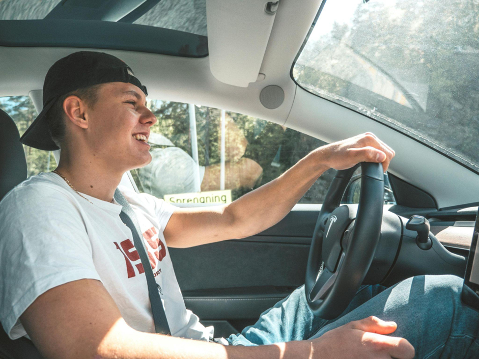 A young man wearing a white t-shirt and backward cap smiles while driving a modern car on a sunny day, showing confidence and comfort behind the wheel.